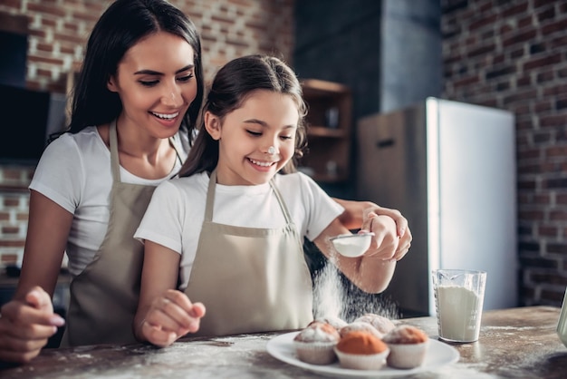 Retrato de mãe e filha sorridentes polvilhando cupcakes cozidos em pó de açúcar na cozinha