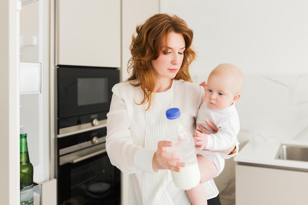 Retrato de mãe pensativa em pé e segurando seu bebezinho com garrafa de leite na mão na cozinha