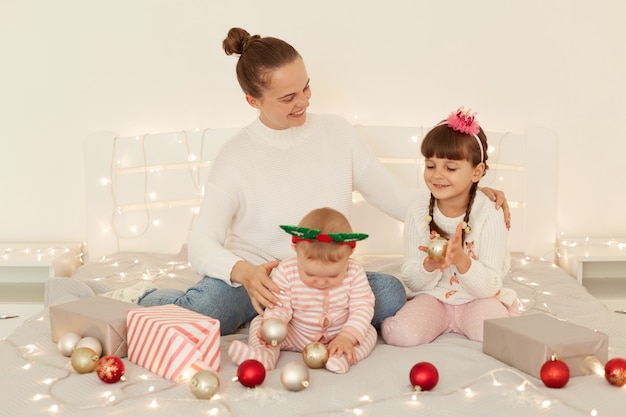 Retrato de mãe sorridente positiva com crianças vestindo trajes casuais, sentados na cama, passando algum tempo juntos durante a véspera de ano novo, comemorando as férias de inverno.