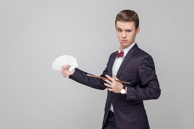 Retrato de mágico elegante com penteado elegante em smoking preto segurando a varinha mágica e apontando para cartas de baralho nas mãos mostrando truque de desempenho ilusionista estúdio tiro fundo cinza