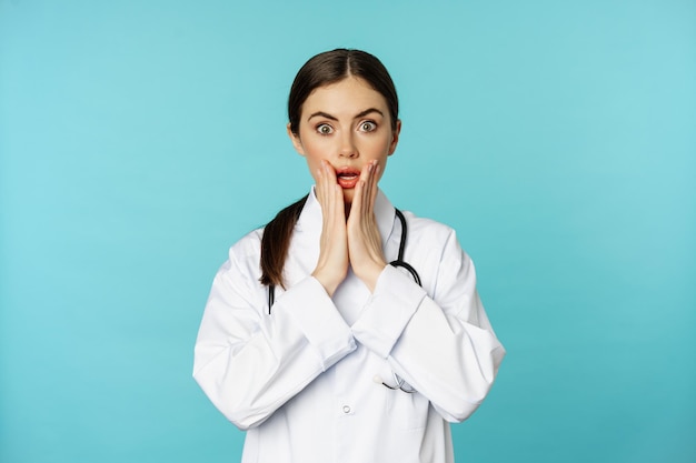 Retrato de médica chocada, estagiária de jaleco branco, olhando preocupado e confuso para a câmera, rosto de descrença, de pé sobre fundo torquesa