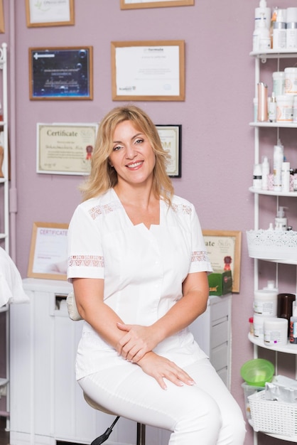 Retrato de médica loira em um salão de beleza de uniforme branco, esteticista olhando para a câmera e sorrindo, médico senta-se em um fundo de certificados.