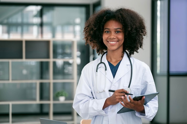 Retrato de médica negra sorridente vestindo jaleco branco com estetoscópio segurando o tablet em pé no consultório do hospital sorrindo para a câmera