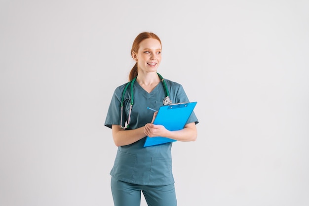 Retrato de médica sorridente em uniforme médico escrevendo prescrição na área de transferência preenche a história médica olhando para longe em pé sobre fundo branco isolado no estúdio