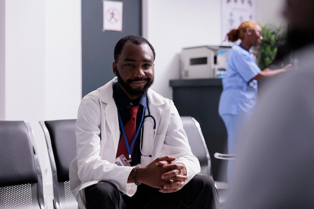 Retrato de médico americano africano no corredor, sentado nos assentos da sala de espera antes de ter consulta médica com os pacientes. clínico geral trabalhando em cuidados de saúde no centro.