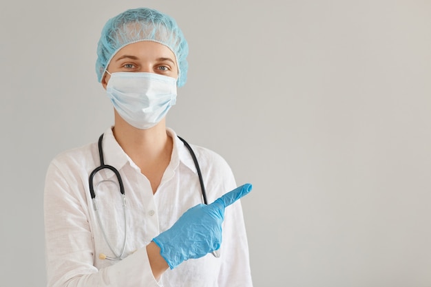 Retrato de médico de mulher otimista usando boné médico, luvas e máscara protetora em pé apontando de lado com o dedo indicador, apresentando espaço de cópia para propaganda.