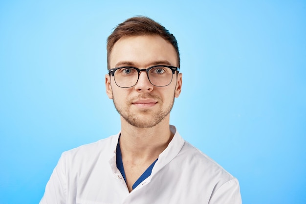 Retrato de médico estagiário de jaleco branco e óculos com uma expressão facial séria isolada no fundo da parede azul