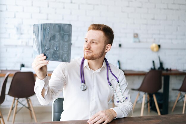 Retrato de médico masculino confiante vestindo uniforme branco examinando tomografia computadorizada do cérebro sentado na mesa no escritório do hospital