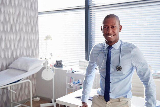 Retrato de médico masculino sorridente com estetoscópio em pé pela mesa no escritório