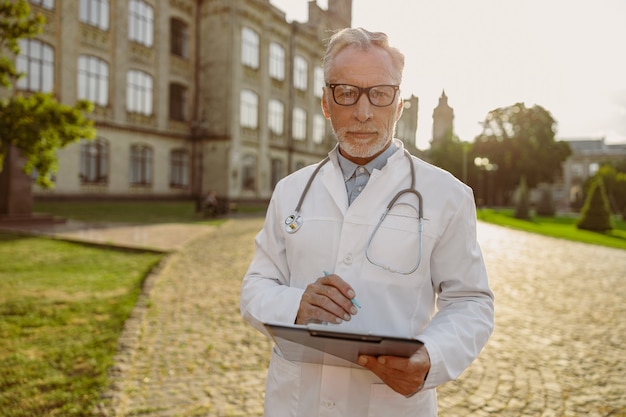 Retrato de médico sênior concentrado, usando jaleco e óculos, segurando uma prancheta, olhando