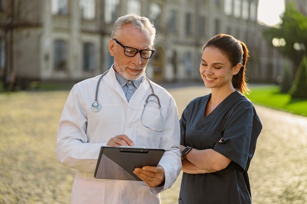 Foto retrato de médico sênior concentrado, usando jaleco e óculos, segurando uma prancheta, olhando