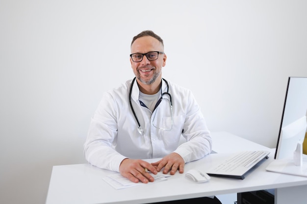 Retrato de médico sorridente em óculos e jaleco branco sentado em seu local de trabalho no escritório