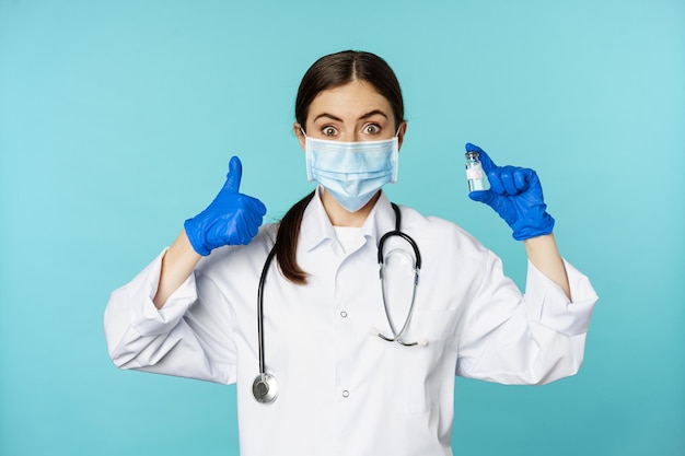 Retrato de médico sorridente, pessoal médico em máscara médica facial e luvas de borracha, mostrando os polegares para cima e coronavírus, vacina omicron, sobre fundo azul.