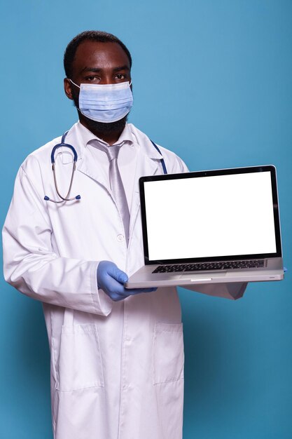 Retrato de médico usando estetoscópio, máscara facial e luvas de látex mostrando laptop com maquete de tela branca para o conceito de saúde. Médico de jaleco segurando computador portátil.