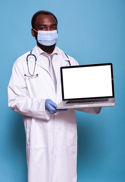 Retrato de médico vestindo jaleco mostrando laptop com maquete de tela branca para o conceito de cuidados de saúde. Médico em equipamentos de proteção médica com estetoscópio segurando um computador portátil.