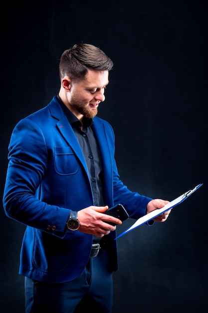 Retrato de meio comprimento de jovem bonito sorridente vestindo camisa escura e terno azul segurando pasta azul olhando para a câmera fundo preto conceito de dinheiro e negócios