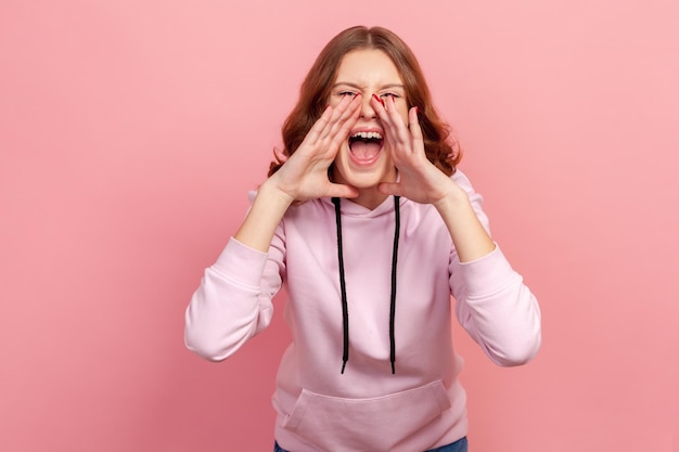 Retrato de menina adolescente agressiva com cabelo encaracolado, gritando anúncio em voz alta, segurando as mãos ao redor da boca gritando notícias loucas Tiro de estúdio interior isolado no fundo rosa