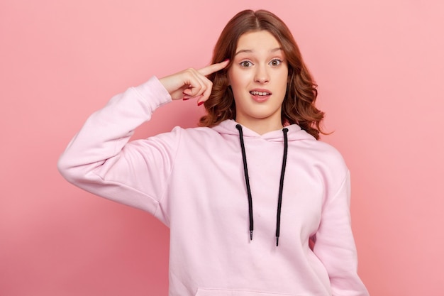 Retrato de menina adolescente com capuz mostrando gesto estúpido com o dedo perto da cabeça, fora da mente, acusando plano idiota louco, expressão imprudente. Tiro de estúdio interior isolado no fundo rosa