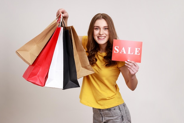 Retrato de menina adolescente de cabelos castanhos sorridente em camiseta amarela segurando sacolas de compras e mostrando a inscrição de venda, feliz com descontos. Tiro de estúdio interior isolado em fundo cinza.