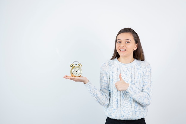 Retrato de menina adolescente em suéter segurando o relógio despertador e dando polegares para cima.