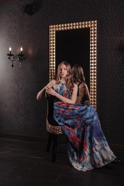 Retrato de menina adolescente muito bonito em um vestido elegante no espelho no interior elegante escuro da sala de estar. Emoções de crianças e poses. Conceito de estilo, moda e beleza. Copie o espaço para o site