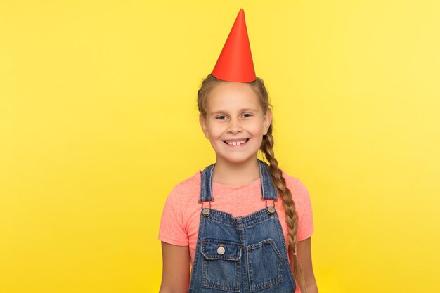 Retrato de menina adorável alegre de macacão jeans com cone engraçado na cabeça sorrindo para a câmera, aproveitando a festa de aniversário, celebração do feriado. tiro de estúdio interno isolado em fundo amarelo