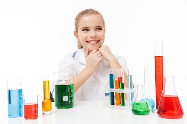 Retrato de menina alegre com jaleco branco fazendo experimentos químicos com líquido multicolorido em tubos de ensaio isolados sobre uma parede branca