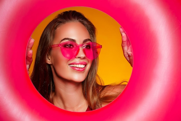Retrato de menina alegre em óculos de sol rosa que segura círculo de borracha sobre fundo de cor amarela