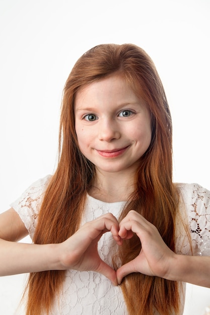 Retrato de menina alegre sorridente, fazendo formato de coração com as mãos no fundo branco.