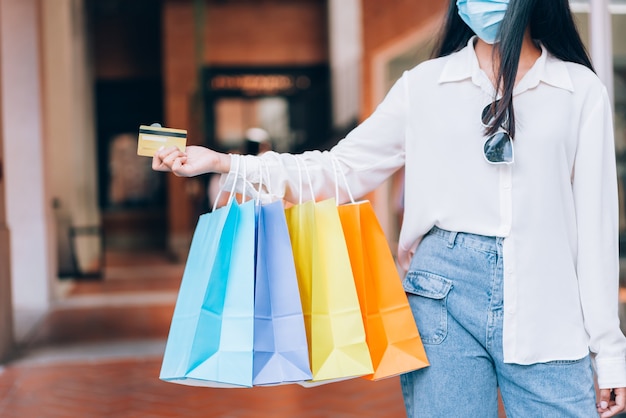 Retrato de menina asiática animado linda garota usando máscara protetora feliz, sorrindo com segurando o cartão de crédito e sacolas de compras, desfrutando em compras de expressão relaxada, conceito de estilo de vida.