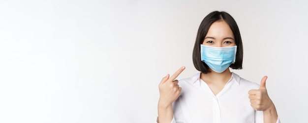 Retrato de menina asiática em máscara médica mostrando os polegares para cima e apontando para sua proteção covid em pé sobre fundo branco