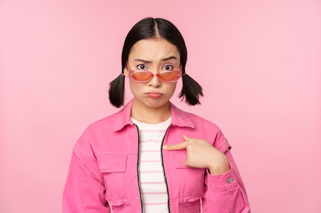 Retrato de menina asiática parece confuso e aponta para si mesma cara perplexa olha com descrença na câmera fica sobre fundo rosa