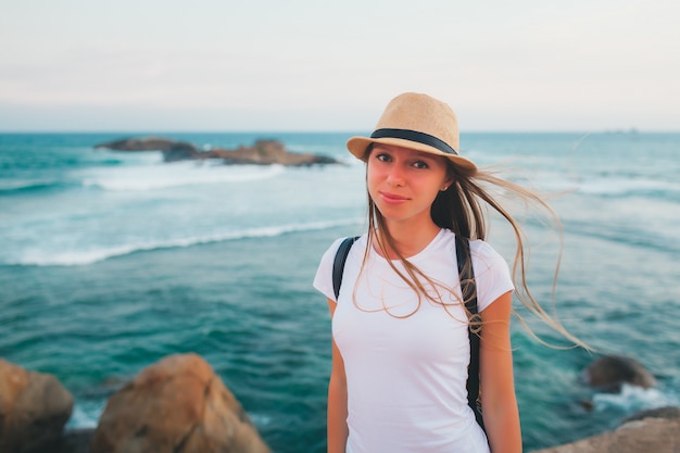 Retrato de menina atraente viajando de chapéu e camiseta branca com mochila na praia do penhasco ao pôr do sol