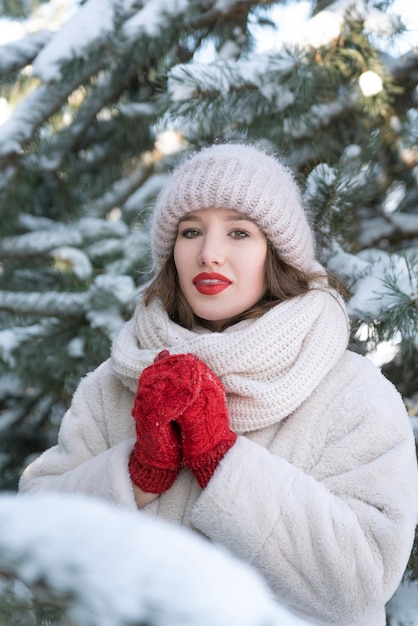 Retrato de menina bonita com casaco de pele branco, chapéu e luvas vermelhas sobre fundo de pinheiros cobertos de neve. Moldura vertical