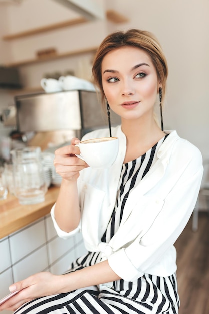 Retrato de menina bonita olhando pensativamente de lado enquanto está sentado no balcão e bebendo café na cafeteria. Senhora bonita com cabelo loiro na camisa branca, sentado no restaurante com o copo na mão