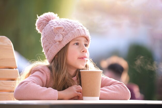 Retrato de menina bonitinha de chapéu rosa sentado sozinho no café da rua bebendo chá de copo de papel