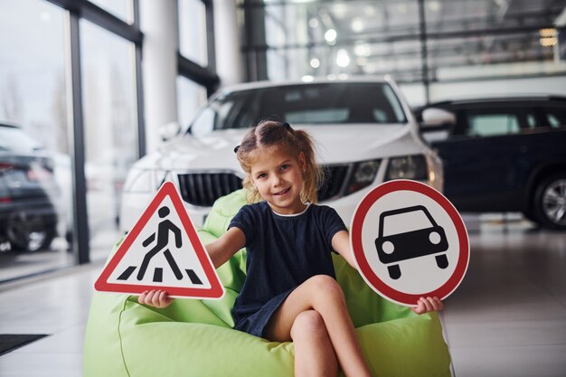 Retrato de menina bonitinha que segura sinais de trânsito nas mãos em salão de automóvel.