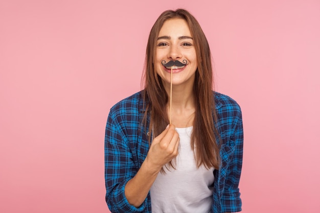 Retrato de menina brincalhão alegre na camisa quadriculada segurando o bigode enrolado falso na vara e sorrindo para a câmera se divertindo usando o estúdio de acessórios de baile de máscaras isolado no fundo rosa