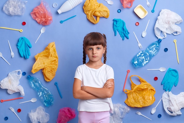 Retrato de menina chateada vestindo camiseta branca posando contra a parede azul e muito lixo plástico ao redor olhando para a câmera com lábios de beicinho tristes com o perigo para a ecologia