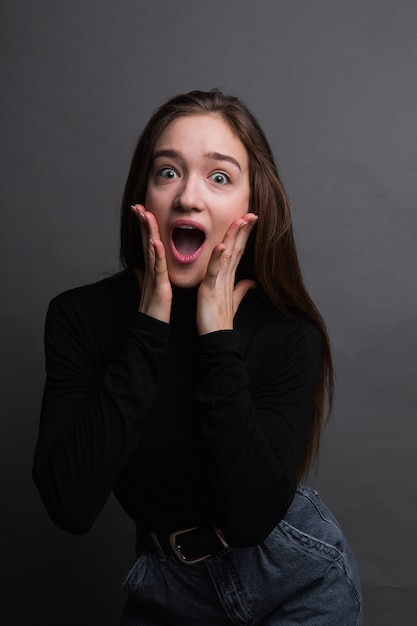 Foto retrato de menina chocada na camisa preta sobre fundo cinzento escuro. jovem surpreendida