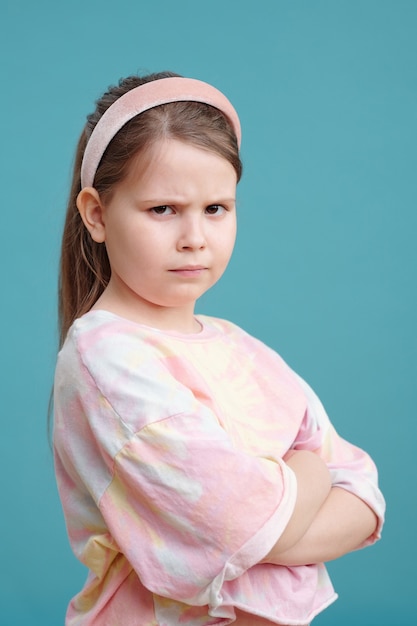 Retrato de menina com cara de zangado em pé com os braços cruzados contra o fundo azul