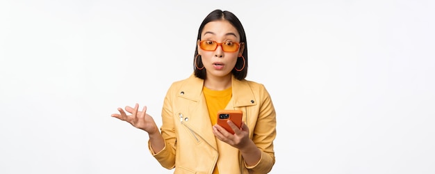 Retrato de menina coreana em óculos de sol segurando smartphone parecendo confuso e encolhendo os ombros em pé sobre fundo branco