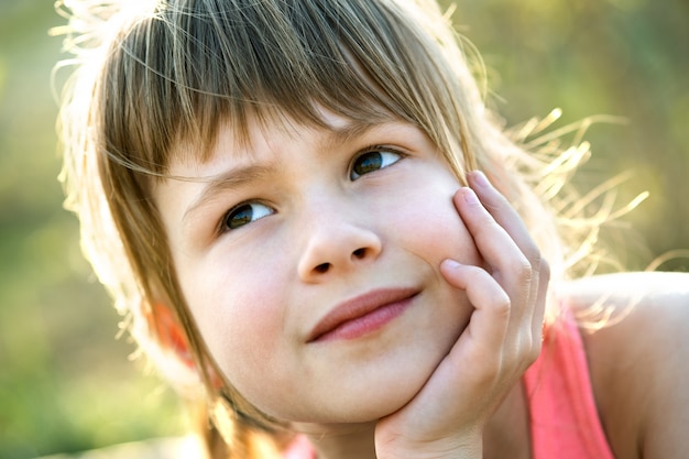 Retrato de menina criança bonita com olhos cinzentos e cabelo longo loiro, apoiando-se nas mãos dela sorrindo alegremente ao ar livre no fundo desfocado brilhante