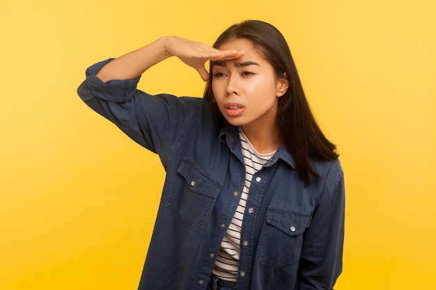 Retrato de menina curiosa na camisa jeans, olhando cuidadosamente para longe com a mão sobre os olhos, escolhendo a distância, explorando o horizonte com vista atenta. tiro de estúdio interno isolado em fundo amarelo