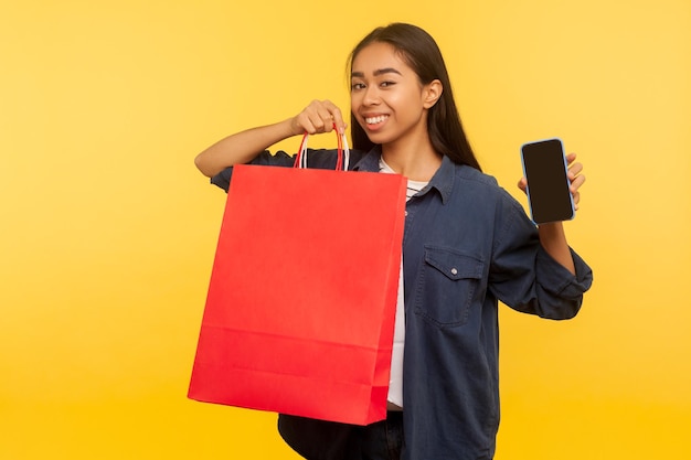 Retrato de menina de cliente feliz em elegante camisa jeans segurando sacos e celular com display vazio simulado, satisfeito com compras na loja de moda, brechó. tiro de estúdio interno isolado