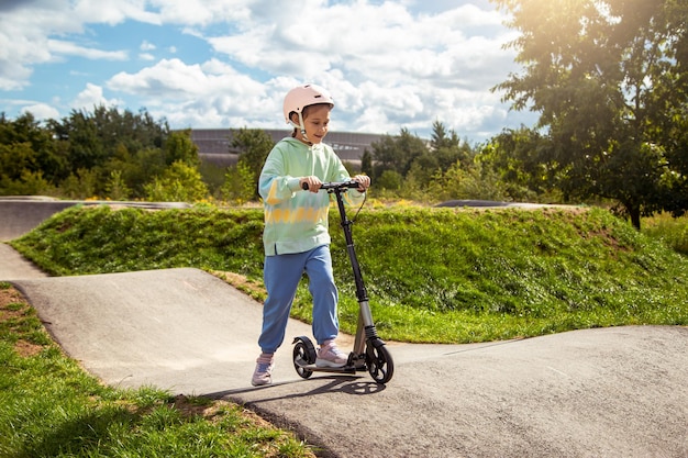 Retrato de menina de escola caucasiana bonitinha usa capacete se diverte andando de scooter no parque de rua asfaltado trackxAin ao ar livre em dia ensolarado Atividades saudáveis para crianças fora