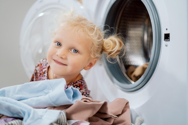 Retrato de menina de olhos azuis com cabelo loiro sorri para a câmera na máquina de lavar aberta de fundo