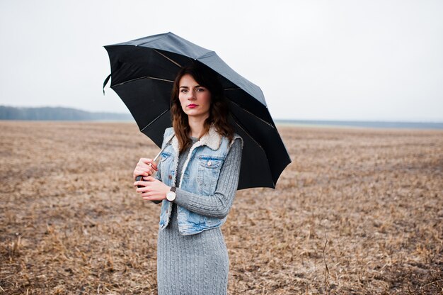 Retrato de menina encaracolado morena em jaqueta jeans com guarda-chuva preta no campo.