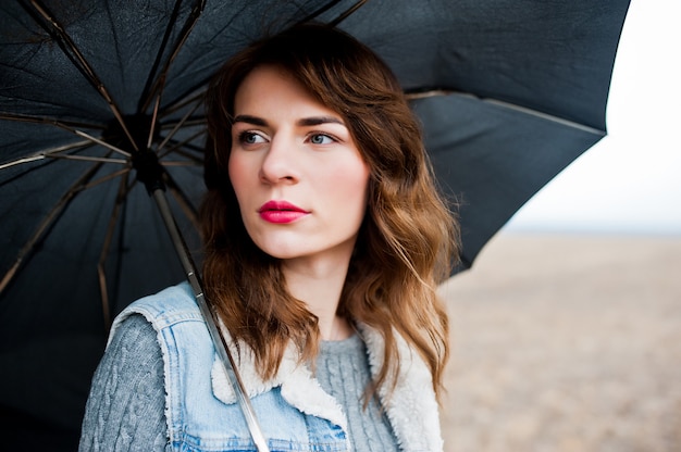 Retrato de menina encaracolado morena em jaqueta jeans com guarda-chuva preta no campo.