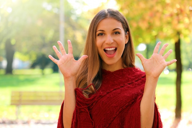 Retrato de menina engraçada positiva rindo vestindo poncho vermelho no parque na temporada de outono.